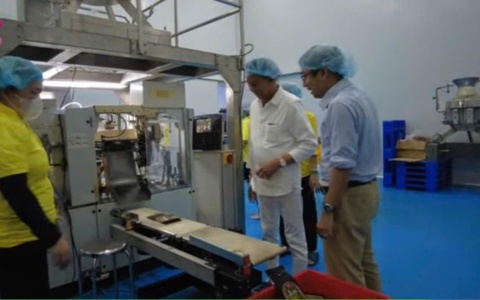 Khách tham quan đội mũ trùm tóc đứng cạnh máy đóng gói trong xưởng